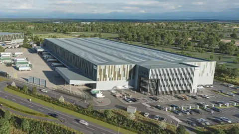 A large, grey industrial unit sits in the middle of green countryside and is surrounded by a road. It has car parking and spaces for lorries to park and load.