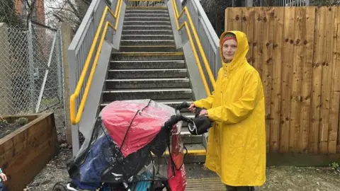 A woman in a yellow coat holding a pushchair. She is stood at the bottom of some metal stairs.