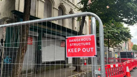 A metal fence with a red sign reading "dangerous structure keep out". The fence is set up outside a derelict building. 