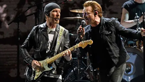 The Edge in black leather jacket and grey hat, playing a guitar, alongside Bono singing into microphone.