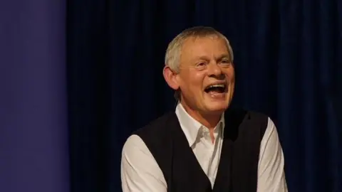 Pete Yendell Martin Clunes laughing with his mouth wide open while sitting on stage at Dorchester Literary Festival. He is wearing a white shirt with an unbuttoned collar and a black waistcoat. He has short grey hair.