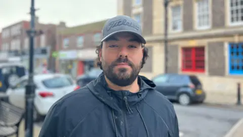 A man in a jacket and cap, looking at the camera. A street lined with cars and buildings is seen in the background.
