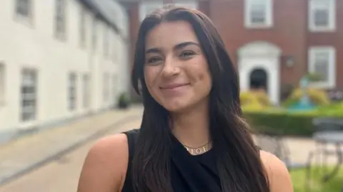 Beth Denman, a woman with long dark hair, is smiling into the camera. She is wearing a gold necklace and a black sleeveless top. 