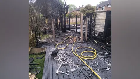 A garden with a severely burned out structure in the centre. Wooden decking in the foreground is severely charred.