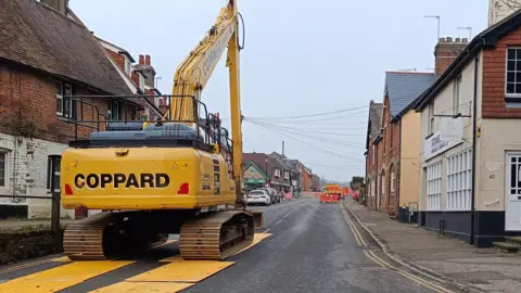 PA Media A yellow digger is parked at the top of a road with houses on either side. 