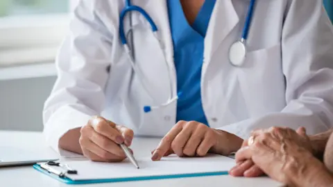 A doctor, wearing scrubs and a white coat, sitting at a desk with a clipboard on it. They are holding a pen. The hands of an older person are also visible in the picture. 