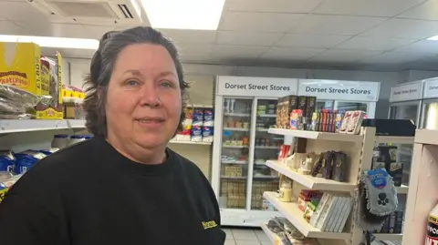 Dawn Venton, a woman with dark hair, wearing a dark, round-necked jumper. She is standing in a supermarket aisle, with fridges in the background. Food is visible on shelves behind her.