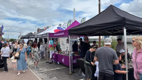 North East Lincolnshire Council Stalls lining the docks in Grimsby. People are walking and gathering at the stalls. The sky is blue with white clouds. 