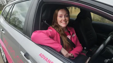 Jenny White is looking out of the driver seat of a car. She is smiling at the camera, has long brown hair and a nose ring. She is wearing a bright pink jacket and t-shirt.