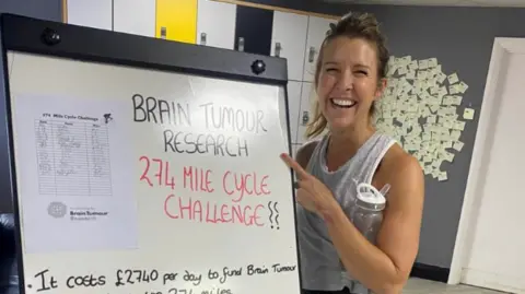 Brain Tumour Research Ms Wrathall smiles at the camera, wearing a grey vest and carrying a water bottle. She is standing beside a movable white board that reads: Brain Tumour Research, 274 Mile Cycle Challenge. 