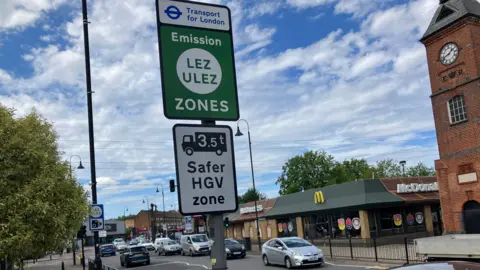 A Transport for London roadside sign marking the LEZ and ULEZ zones is mounted on a pole beside a busy road. Cars are travelling through a junction and a McDonald's restaurant and brick clock tower can be seen in the background under a partly cloudy sky.