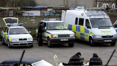 Getty Images Police cars and vans are parked at an industrial estate in Welling after bagged-up stashes of notes were found there. An officer is standing by a vehicle talking to police inside it. Two people are in the foreground, working.