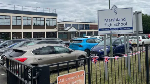 John Devine/BBC A gate leads into a school car park where several vehicles are parked. In the background is a school which has two storeys and large windows.