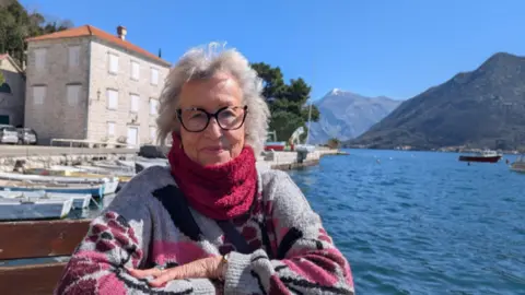 Anita in a midshot wearing a fluffy jumper and red snood, smiling to camera with an exotic location behind with a river and mountains in the background. There are moored rowing boats just behind her and a three storey building