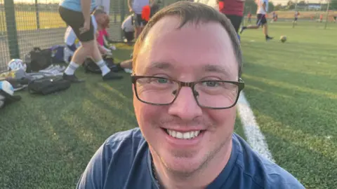 A person in a blue sports top stands beside an outdoor football pitch while a game takes place in the background.