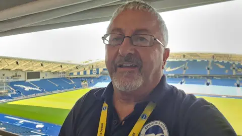 Nigel Summers A man with a white goatee and short white hair standing in The Amex stadium. He is wearing a navy polo shirt with the Brighton & Hove Albion badge on it.