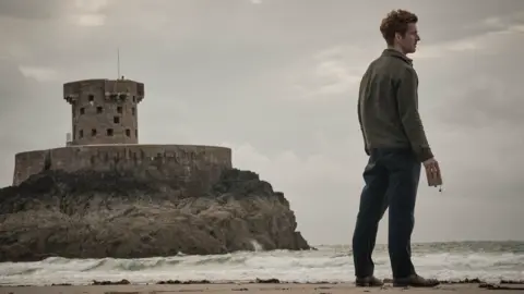 UKTV Damien Molony is holding an empty alcohol flask looking out onto the ocean. He's standing on St Ouen's Bay with La Rocco tower and a moody, grey sky behind him.