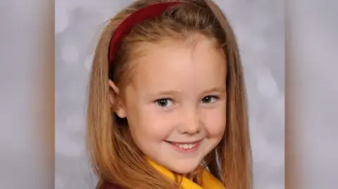 A headshot of Elsie Dot Stancombe. She had long brown hair, tied back with a red headband, and is smiling.