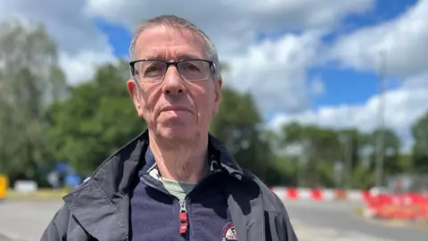 Julia Gregory / BBC A man, Bob Oliver, wearing a zip up fleece and coat. He is stood in front of a road with roadworks next to it.