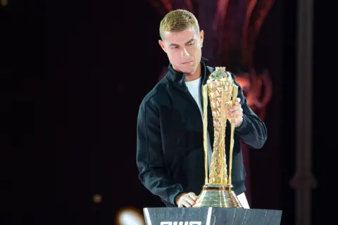 Esports Foundation Cristiano Ronaldo, sporting a blonde dyed hairstyle and wearing a black Harrington jacket over a white t-shirt, carefully places the tall, golden EWC trophy on top of a dark grey plinth.