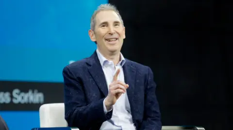 Getty Images Amazon CEO Andy Jassy sits and gestures with his hands as he speaks, wearing a light-speckled navy blue jacket and light blue shirt.