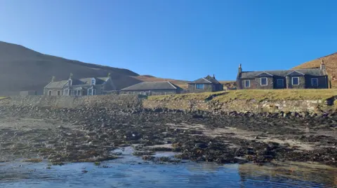 Mowi A group of cottages on a shoreline of Sanda. There is a rocky beach and a blue sky above the island.