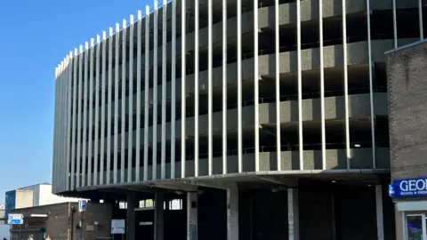 Exterior shot of the George Street car park in Hull. It is a 1960s Brutalist-style structure. Access to the car park has been blocked off with red traffic barriers. There is a person seated on the wall at the front of the facility.