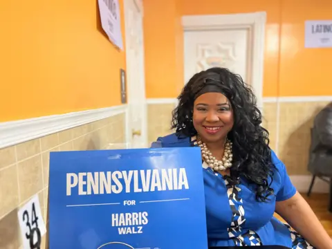 Kari Holmes, wearing a blue shirt, smiles while holding a Pennsylvania for Harris Walz sign