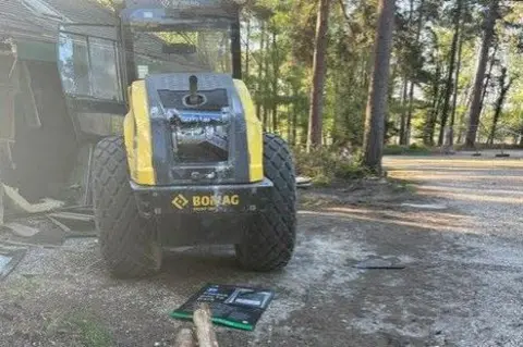 Forestry England A close up of the construction vehicle. It is yellow in colour and it has very large wheels. It has smashed windows and other marks on its bodywork. A sign is on the ground, likely knocked over by the vehicle.
