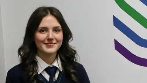 Shimna Integrated College A girl with dark hair is standing in front of a white wall with her school logo to her right. She is wearing a navy blue school uniform.