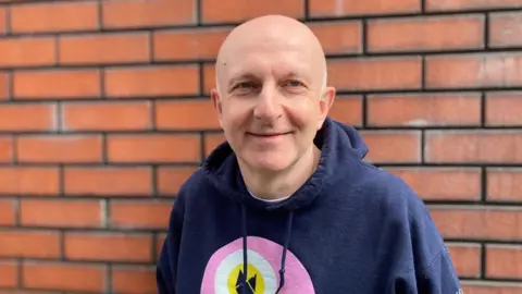 A bald man smiles at the camera. He is wearing a dark navy hoodie, with a pink, white, and yellow circle in the middle. He is standing in front of a red-brick wall.