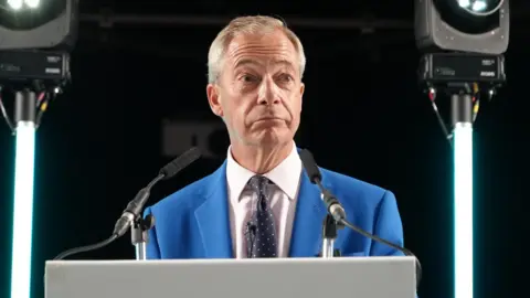 BBC Nigel Farage standing at a rally wearing a bright blue suit and polka dot tie looking out into the crowd. 