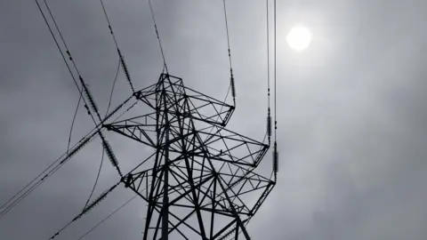 An electricity tower pictured silhouetted against a grey sky in Inverness. The structure is tall with metal lattice work and cables attached to it. 