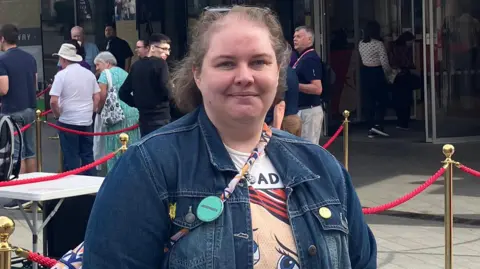 A woman with short brown hair wearing a denim jacket stands outside the Crucible as people queue up to enter for the day