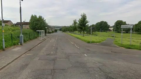 An empty suburban road with park on thr right. There is a bus stop on the left, many trees, and a sign saying Auchenbank Community Park on the right.