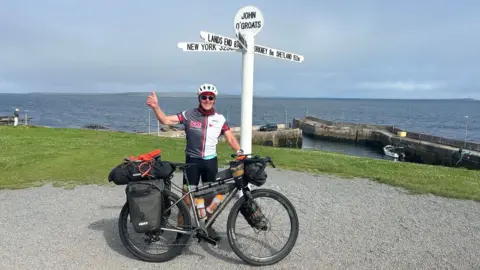 Norman Brown with his one thumb up wearing sunglasses and a helmet holding his bike next to the John O'Groats landmark. The sea is in the background.