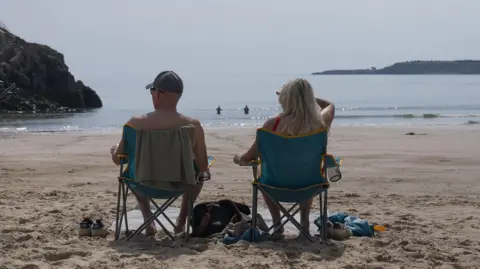 The backs of two people sat on deckchairs. One is a topless man wearing a baseball cap and sunglasses with a towel on the back of his blue chair. The other is a woman with long blonde hair in a red swimsuit, sat in a matching blue chair with one hand up to shield her eyes from the sun. The beach is sandy, and they have bags and shoes beside them and are looking out at the sea where two more people are swimming. There are cliffs on either side. 