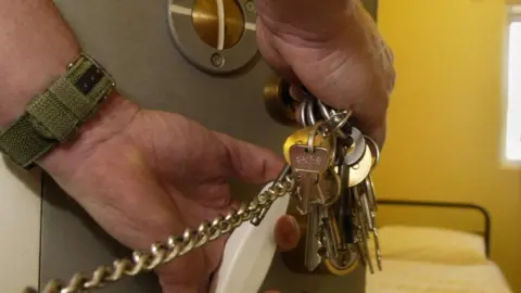 A man's hands with a large set of keys on a chain with one in a keyhold. A prison bed is in the cell beyond.