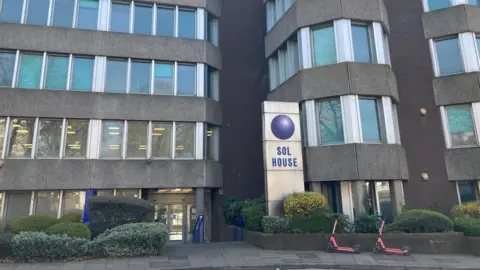 A block of flats with lots of glass windows and a dirt pillar with "Sol House" written on it.