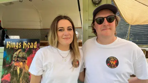 A head and shoulders image of a woman and a man wearing white T-shirts with the King's Head logo on them. They are standing in front of a mobile bar and next to a large poster of The King's Head logo, which depicts a Viking-type character under the words King's Head.