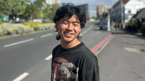 Family handout Yubin Tamang wearing a black t-shirt with a pattern stands on the side of the road and smiles. He has dark hair.