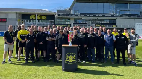 Gloucestershire CCC Players and staff of Gloucestershire County Cricket Club stand on the pitch at the county ground in Bristol in a team formation. In front of them is a plinth with the T20 Blast trophy on it. It is a sunny day and the main stand at the stadium is visible behind them