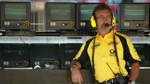 A man with brown and grey hair stands in front of multiple TV screens. He is wearing bright yellow head phones with a microphone attached, a walkie talkie and a yellow polo shirt with some branding on. 