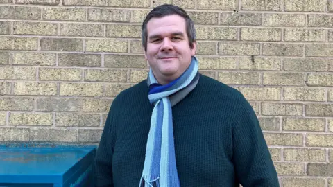 A man with short dark hair, a blue scarf and dark jumper, stands and smiles in front of a brick wall.