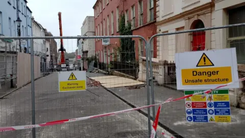BBC Shows a barrier with a sign saying "Danger - unsafe structure" and buildings on either side of the street and a crane in the background