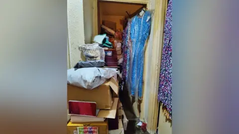 The hallway of a house, filled with carboard boxes, bags and other items which are piled on the floor. Dresses are also hung on a door.