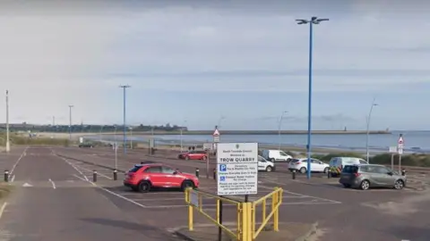 The entrance of Trow Quarry car park in South Shields. There is a sign explaining the parking payment and rules, with a number of vehicles parked at the site. The se and the beach are nearby, with Tynemouth Priory and Castle visible further in the distance. 