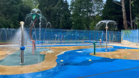 An area of the pool with water features including mushrooms and cannons. The water is bright blue and there are areas that are sandy coloured and made to look like a beach.