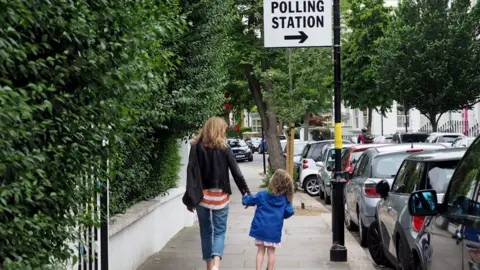 A woman and a young child walking down a tree-lined suburban street hand in hand. Attached to a lamppost is a sign that says 'polling station'.