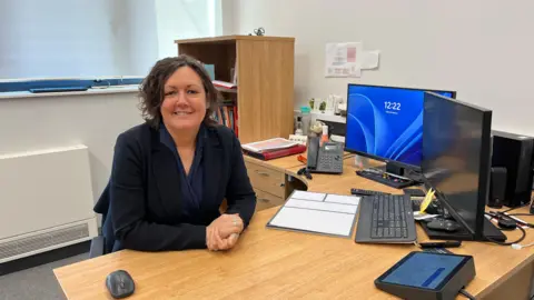 Photograph of Phillipa Lewis in a smart suit sitting behind a desk with two computers on her desk.